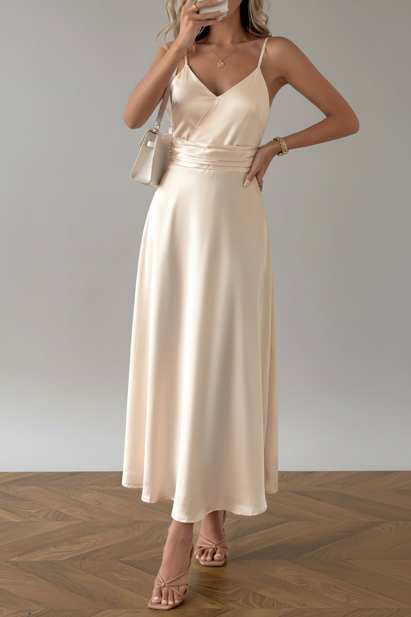 Woman in a beige dress standing in a room with white curtains and a chair.