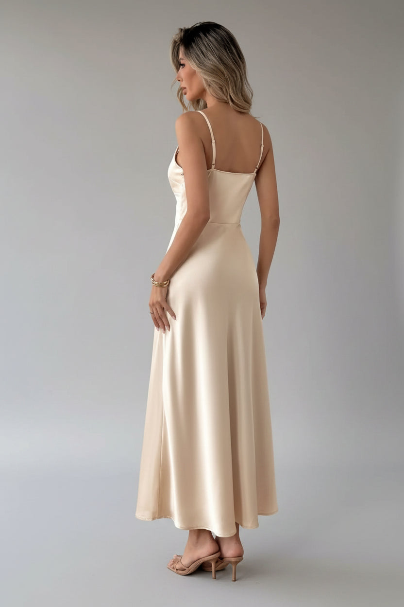 Woman in a beige evening gown standing in a room with white curtains and a chair.