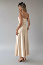 Woman in a beige evening gown standing in a room with white curtains and a chair.