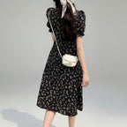Woman in a black floral dress standing on grass with a white bag.