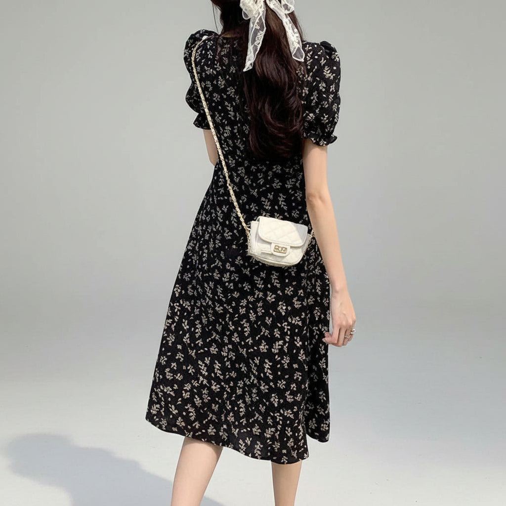 Woman in a black floral dress standing on grass with a white bag.