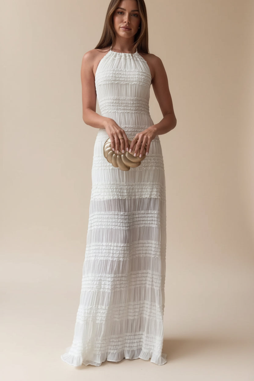 Woman in a white halter neck dress holding a gold clutch in a room with shelves.