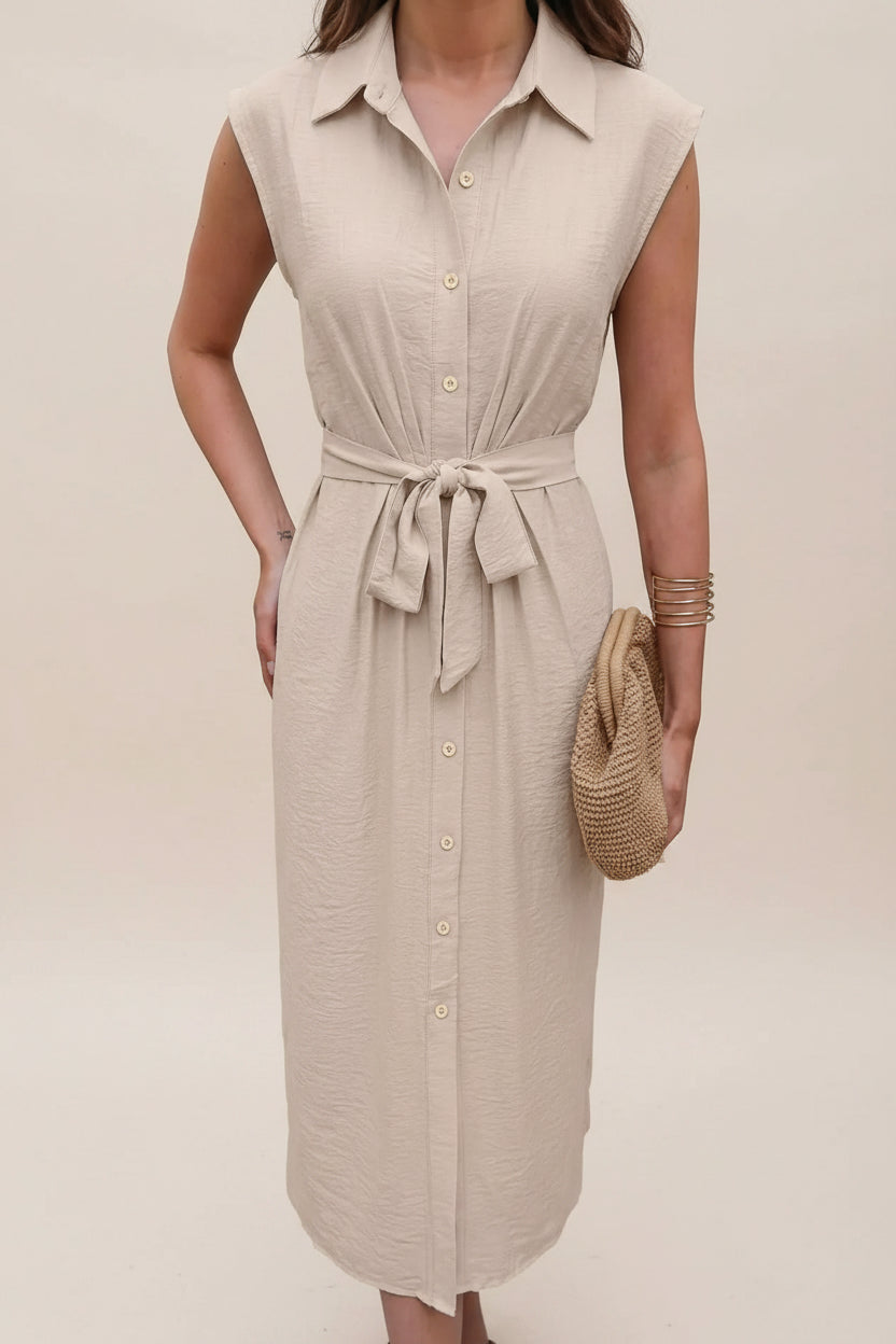 Woman wearing a beige sleeveless dress with a belt, holding a straw bag indoors.