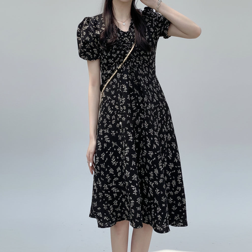 Woman wearing a black floral dress standing outdoors with flowers and water in the background
